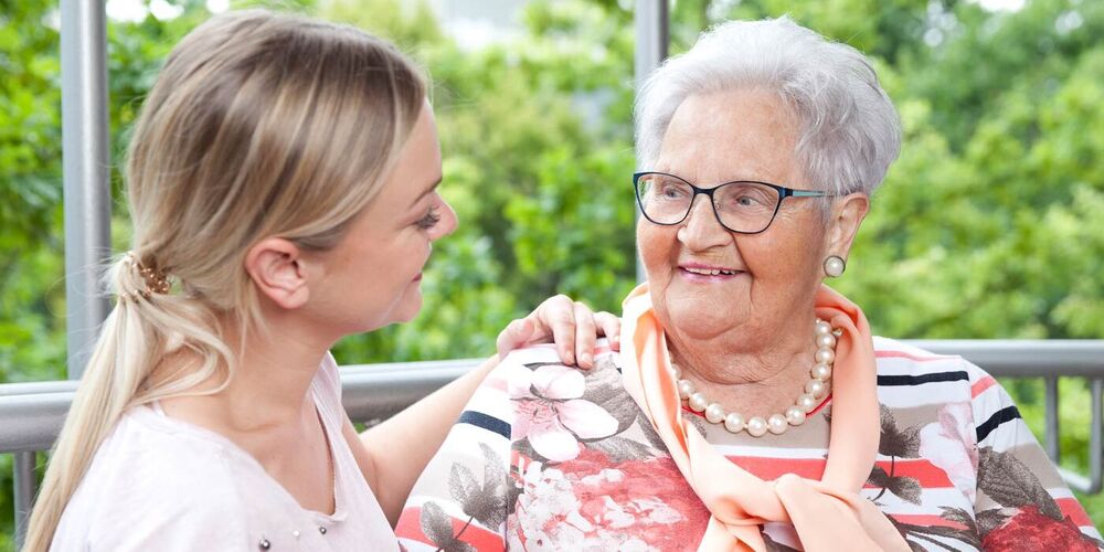Eine Seniorin und eine junge Frau schauen sich an. Sie halten eine Hand und die junge Frau hat ihre andere Hand auf der Schulter der Seniorin.
